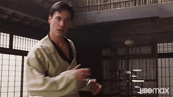 Movie gif. Keanu Reeves as Neo in The Matrix inside of a dojo hopping and smiling and loosening up in preparation to fight.