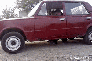 Video gif. Footage of a red car "driving" past us. The car clearly has a hole in the bottom, and the driver and his passenger are moving the car with their feet like the Flintstones.