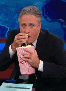 TV gif. John Stewart from the Daily Show leans over his desk with an intense, almost zombie-like state. He shovels popcorn, almost missing his mouth because he’s so absorbed by what he’s looking at. 