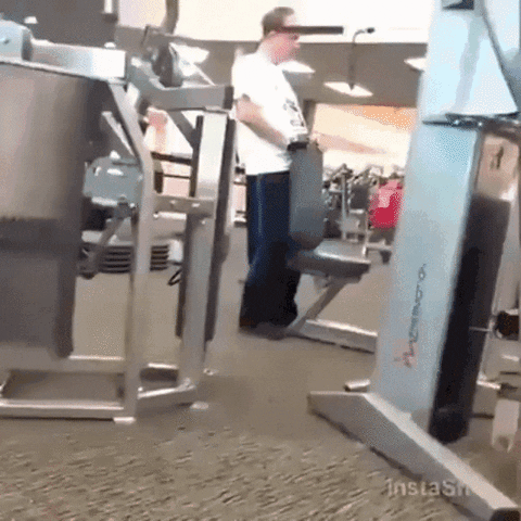 Video gif. Footage of a man at a gym, pulling one of the machines' weighted ropes with his head.