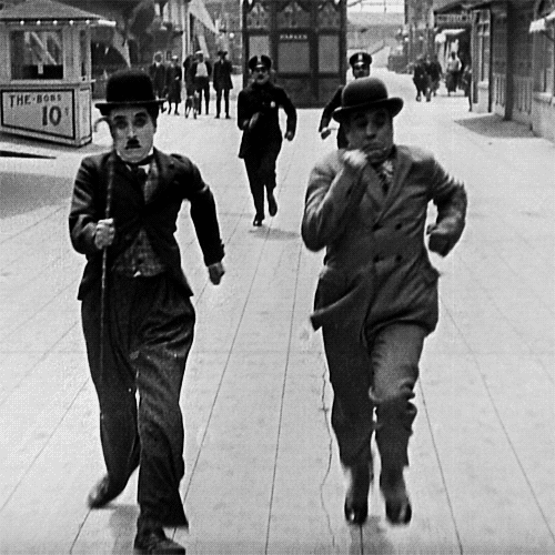 Movie gif. Charlie Chaplin and a friend from The Little Tramp are running away quickly, looking back at the policemen chasing them through the streets.