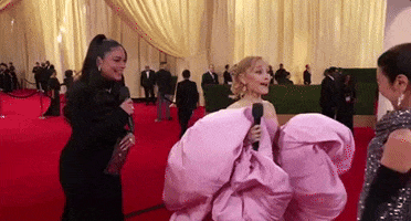 Oscars 2024 GIF. Ariana Grande, dressed in an oversized pink gown, excitedly shuffles up to and greets Michelle Yeoh with a cheek-to-cheek hug as Vanessa Hudgens looks on and smiles.
