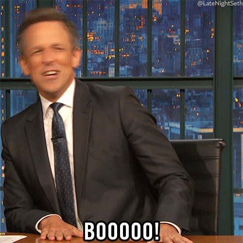 TV gif. Seth Meyers as host of Late Night sits at his desk and wiggles around in his desk chair, looking around, mocking the audience. He shouts out, “Booooo!” 
