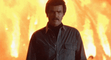 Movie gif. Burt Reynolds as Mex in Heat walks confidently toward us, away from a fiery explosion.