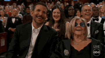Oscars 2024 GIF. Bradley Cooper and his mother Gloria Campano seated at the Oscars, Gloria throwing her head back in laughter and Bradley catching a friend’s eye to laugh with them.