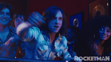 Movie gif. Jamie Bell as Bernie in Rocketman. He closes his mouth and looks at the stage in joy, pointing a finger out in great pride and acknowledgement.