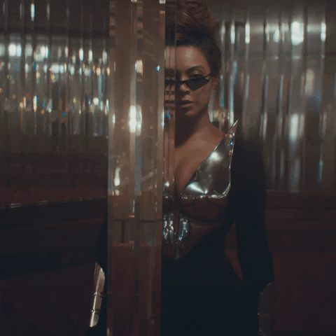 Music video gif. Beyonce gives a questioning glance over narrow sunglasses, peering around a mirrored wall. She's wearing a black dress with a chrome chest plate and bracelets.
