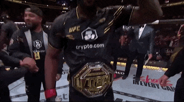 Sports gif. Leon Edwards, a UFC fighter, walks across the ring, making a bicep and pointing at us. Joe Rogan is standing next to him with a microphone as others fill the ring behind him. 