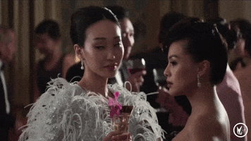 Movie gif. Gemma Chan as Astrid Leong and Victoria Loke as Fiona Cheng from Crazy Rich Asians are wearing very fancy dresses and are holding cocktail glasses in their hands. Their eyes flit up and down and their lips are pursed as they judge everyone who passes by.
