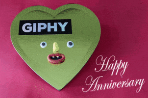 Text gif. The words “Happy Anniversary” sit adjacent to a heart-shaped box labeled “Giphy.” Hands come into frame and remove the lid, revealing bizarre little chocolates with goofy faces on them. 