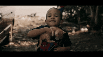 Music video gif. From John Legend's Love Me Now video, a little boy smiles wide while gripping an orange striped kitten to his chest.