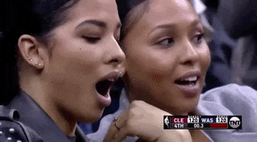 Sports gif. Two basketball fans are sitting courtside at a basketball game and everyone around them is going crazy, standing up and cheering. These two are shell-shocked and sitting in their seat with their mouth agape and arms crossed, unable to comprehend what just happened. 