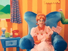 TV gif. Character in Happy Place wearing a 50s style orange wig and an orange and white dress holds a small American flag on a stick as she smiles sweetly, seated on a blue sofa chair on a set decorated in a bright cartoonish style. Text, "Happy Fourth of July."