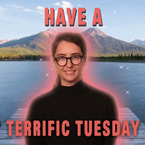 Digital art gif. Studious-looking woman in a black turtleneck and horn-rimmed glasses, glowing with animated sparkles and superimposed over a scenic lake and mountain background, says "Have a terrific Tuesday."