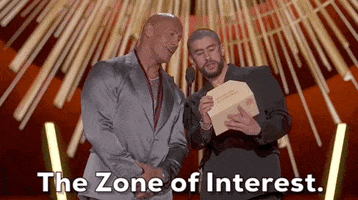 Oscars 2024 GIF. Bad Bunny and The Rock are presenting the award for Best International Feature. Bad Bunny is holding the envelope and he says, "Zone of Interest," and looks midway at the The Rock while he announces, prompting The Rock to say, "Interest" in sync. 