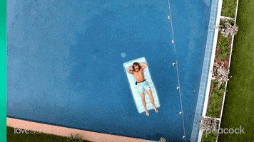 Reality TV gif. Man from Love Island is laying in a pool floaty and a drone camera zooms in on him from above as he lowers his sunglasses and flashes a smile.