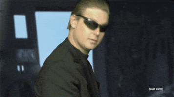 TV gif. Tim Heidecker as Agent Jack Decker from Decker has his hair slicked back and dons an all-black fit with black shades. He nods at us and gives us a cool smirk.