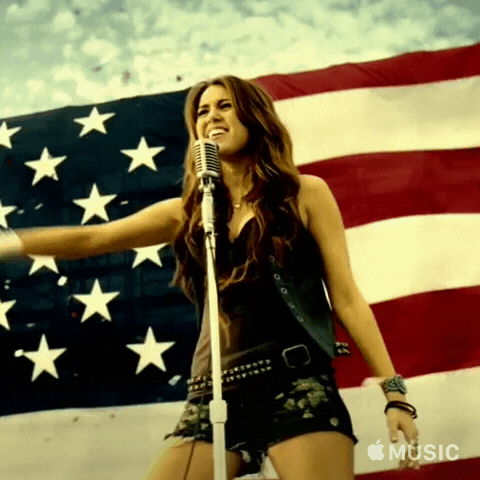 Music Video gif. Miley Cyrus is dancing and singing on stage for her music video for Party In The USA. A giant American flag flutters behind her.