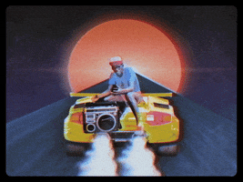 Video gif. Man sits on the back of a speeding sports car with a retro boombox in his hand. The sports car races down a road towards a red sun.