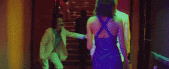 Music video gif. From the video for Neon Indian's Slumlord Rising, a man in a white suit stands at the foot of a sparkling staircase and waves his hand, welcoming a man and woman approaching the staircase.