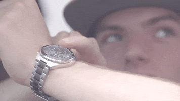 Video gif. A man taps his wrist watch to emphasize haste. 