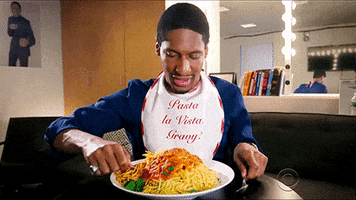 Celebrity gif. Jon Batiste on The Late Show sits in his dressing room with a bib that says, “Pasta la vista, gravy!” He has a huge heaping pile of spaghetti in front of him and he takes two forks and lifts up a massive serving up to his mouth. 