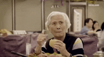 Video gif. An elderly woman holds a hamburger in her hand as she looks over and gives a sincere smile and thumbs up. 