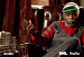 SNL gif. Jay Pharoah wears a tracksuit and a gray mustache as he sits in a tufted leather armchair and gestures to a pile of money on a table beside him.