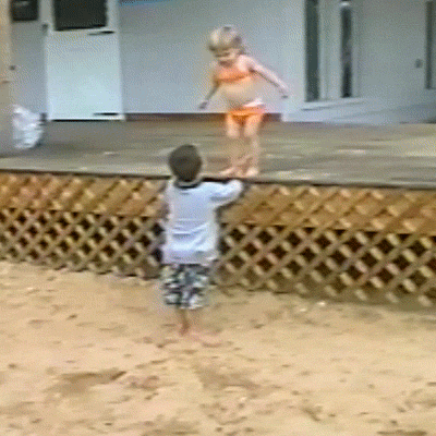 Video gif. Toddler in a bikini jumps off a low deck toward her brother, who attempts to catch her but falls backward into the sand in an adorable fail.