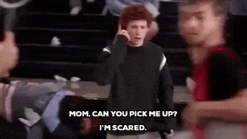 Movie gif. Student from Mean Girls is standing in the middle of the auditorium as chaos ensues around him. He looks incredibly frightened and has called his mom on the phone, asking, "Mom, can you pick me up? I'm scared."