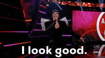 Reality TV gif. Cardi B on the American Music Awards wears a formal black dress holds a microphone and gestures to herself saying, "I look good."