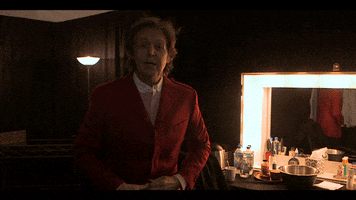 Celebrity gif. Paul Mccartney stands in his dressing room while looking at us. He points and then claps his hands together as he says, “Let’s go!” He then rushes his hand towards us to cover the camera for a cool transition. 