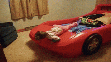Video gif. Toddler lays belly-down on the hood of a racecar-shaped bed, then tumbles forward when another child pushes up the hood from under it.