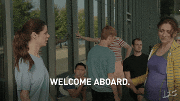 TV gif. Two women on Baroness Von Sketch stand with a small crowd of people. One of them looks tired and shakes her head while she says, “Welcome aboard.” The two women high five. 
