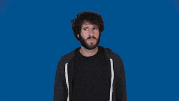 Celebrity gif. Lil Dicky stands in front of a solid blue background, looking from side to side with a slightly confused frown. He puts a hand to his chest as he speaks. Text, "Who me? He grins."