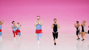 Video gif. Two Groups of people working out together jog into place. One group wears 1980s workout clothes while the other wears more modern sleek outfits. They all clap as they run in place. They then snap into a power stance and point at us. 