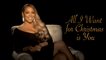 Celebrity gif. Mariah Carey lounges on a white chair, wearing a black dress and sparkling earrings. She smiles and says, “All I want for Christmas is you.”
