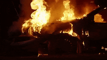 Video gif. Dilapidated car is on fire at night.