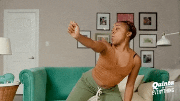 TV gif. Quinta Brunson in Quinta vs. Everything twerks in an orange tank and army green sweats in front of her emerald green sofa at home.