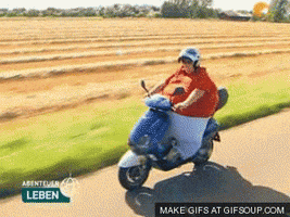 Video gif. A large person zooms by on a motorized scooter, passing fields of crops and their shirt billows in the wind.