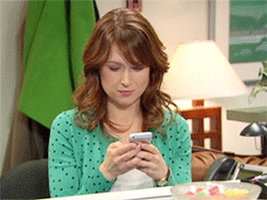 The Office gif. Ellie Kemper as Erin sits at her desk, looking at her phone and laughing.