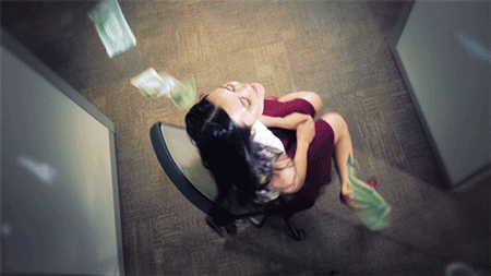 Video gif. From above, we see a woman is spinning on an office chair as she looks up with a smile and multiple dollar bills fall from the sky onto her face.