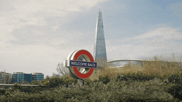 Video gif. London Tube sign reads “Welcome Back” and is surrounded by green bushes, the London Shard building towering in the background.