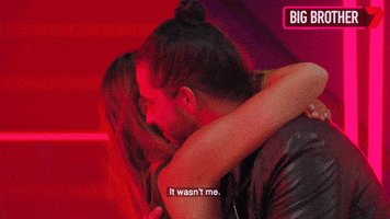 Reality TV gif. We see a man and woman on Big Brother from the side. They have their arms wrapped around each other in a hug as he whispers into her ear, "It wasn't me."