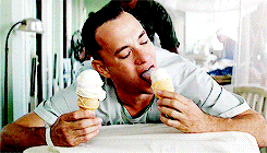 A white man lying down on a bed, licks two ice creams in his hands. 