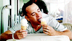A white man lying on a bed, licking two ice cream in his hands.