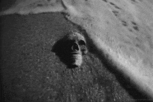 Video gif. A black and white shot of a human skull planted in the shoreline. Waves wash over it and receed, over and over again.