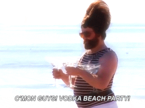 Muž s parukou nesoucí čtyři drinky a jdoucí podél moře s nápisem Vodka beach party.