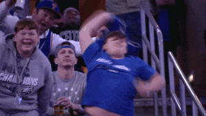 Sports gif. An enthusiastic kid dances while whipping a blue Celtics shirt in the air like a helicopter propeller. Other excited fans cheer on the bleachers behind him.  