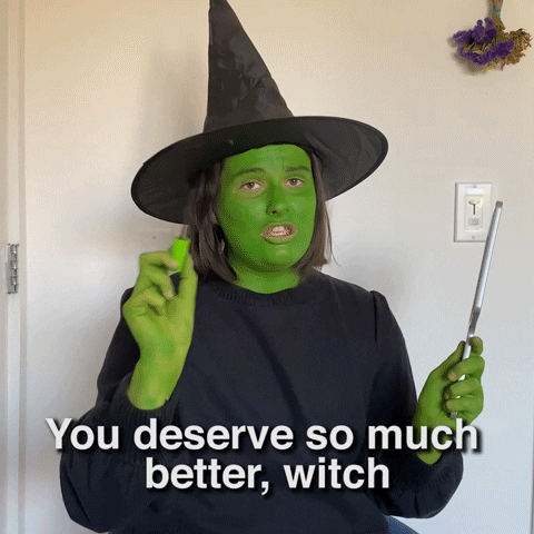 Video gif. Girl wearing a black pointed witch hat and her skin painted green looks at us and speaks before returning to focus on her mascara with a handheld mirror. Text, "You deserve so much better, witch."